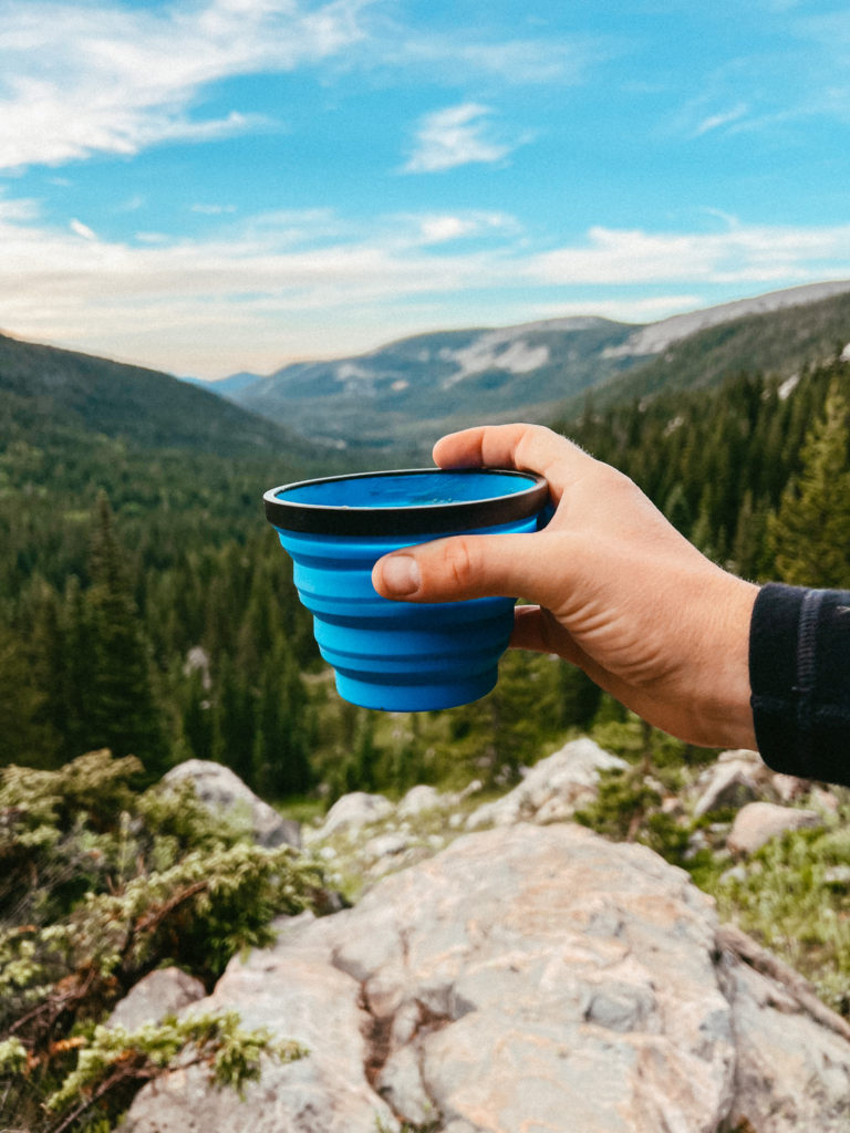 A cup of coffee on a hike with mountains in the distance.