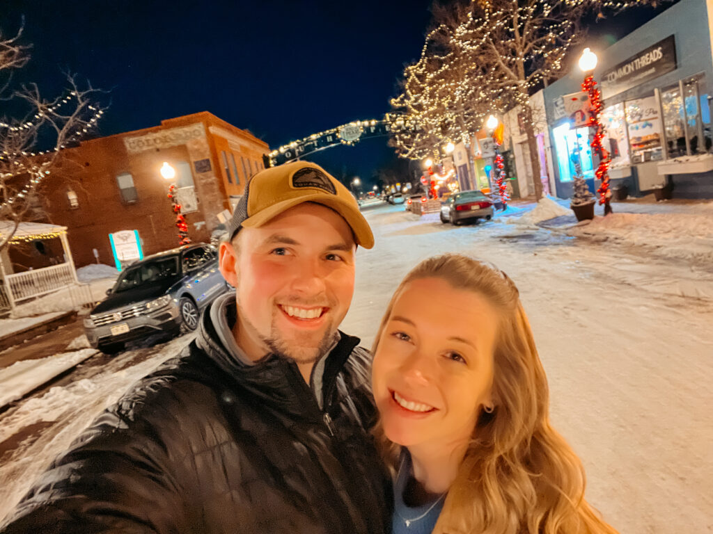 Sam and Abby smiling with snow covered streets and lit up trees in the background.