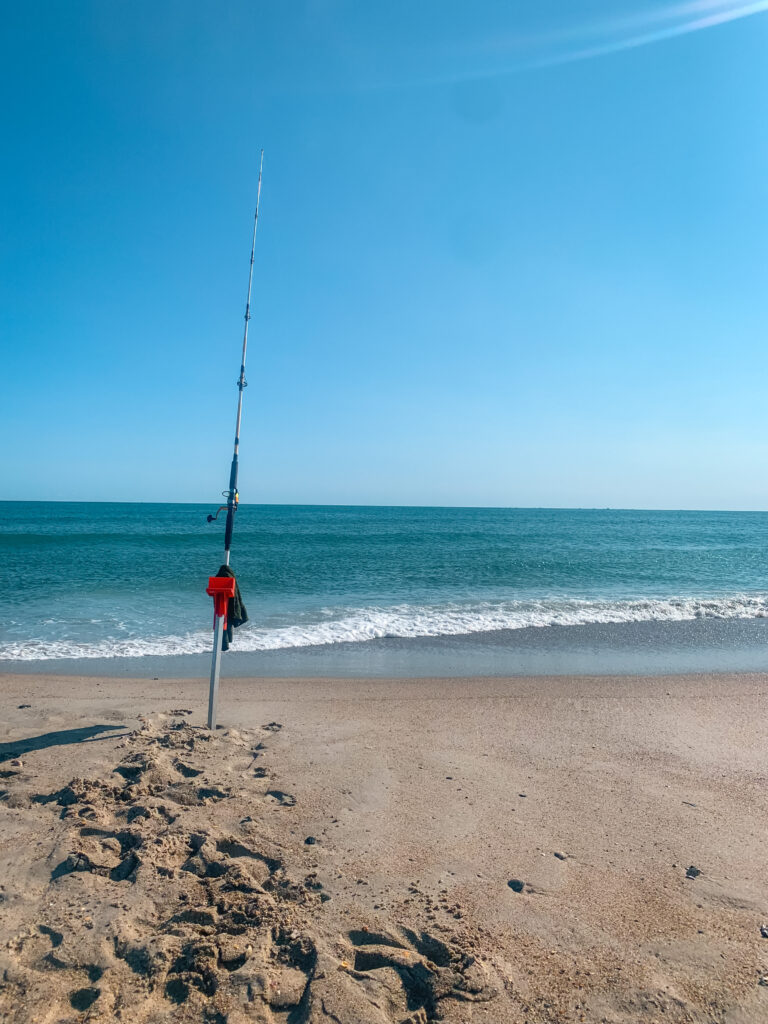 A surf fishing rod in its holder on the beach with ocean waves crashing on the shore.