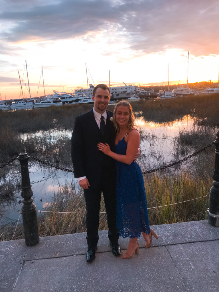 Abby and Sam smiling with sailboats and the sunset in the distance in Charleston, South Carolina.