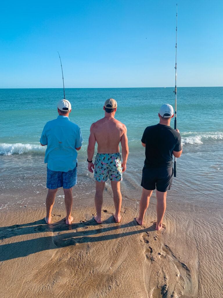 Three men surf fishing on the beach with the ocean and blue skies in the background.