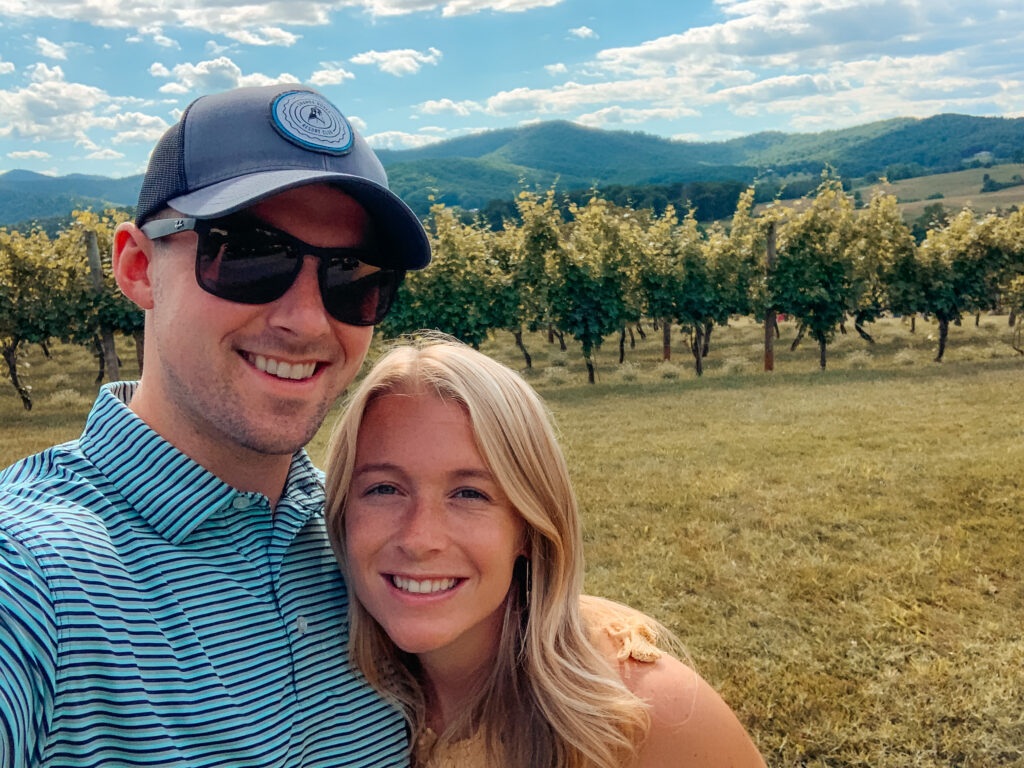 Abby and Sam smiling with a vineyard and mountain views in the background at one of the best wineries in Charlottesville. 
