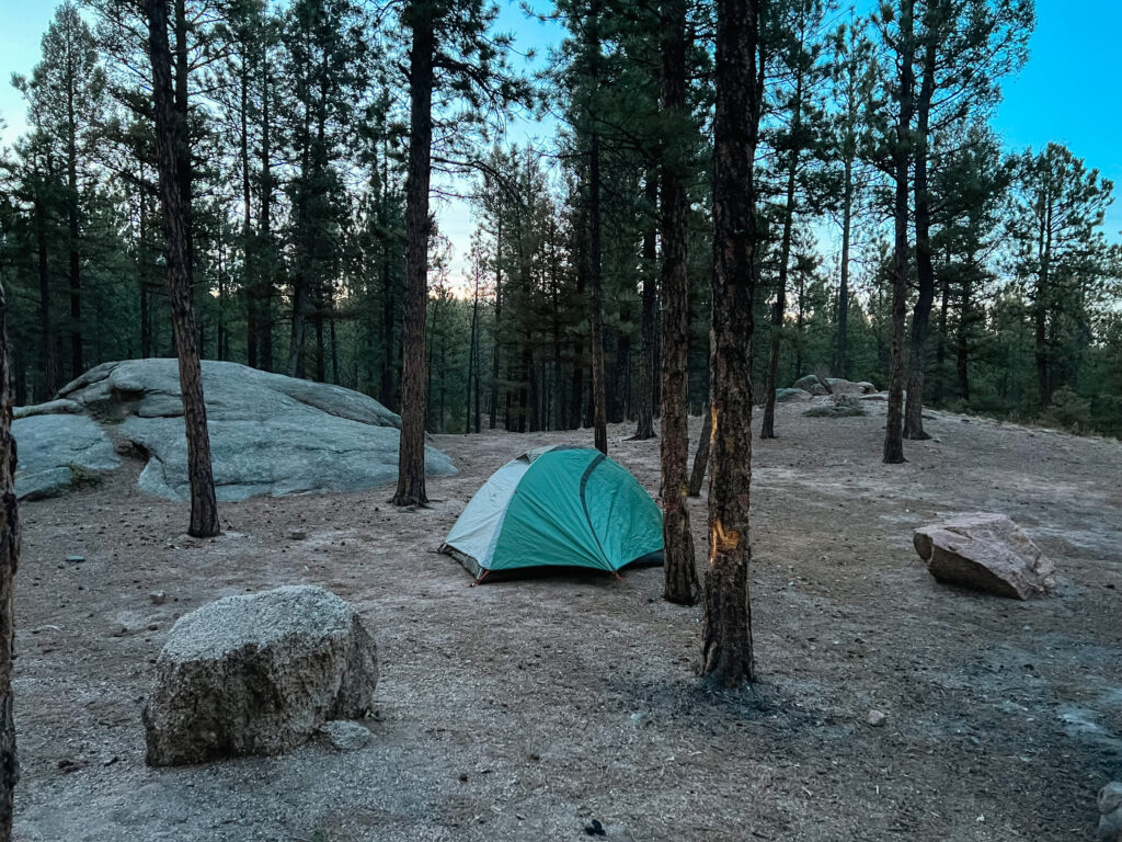 A tent set up surrounded by rocks and trees as the sun sets in the distance.