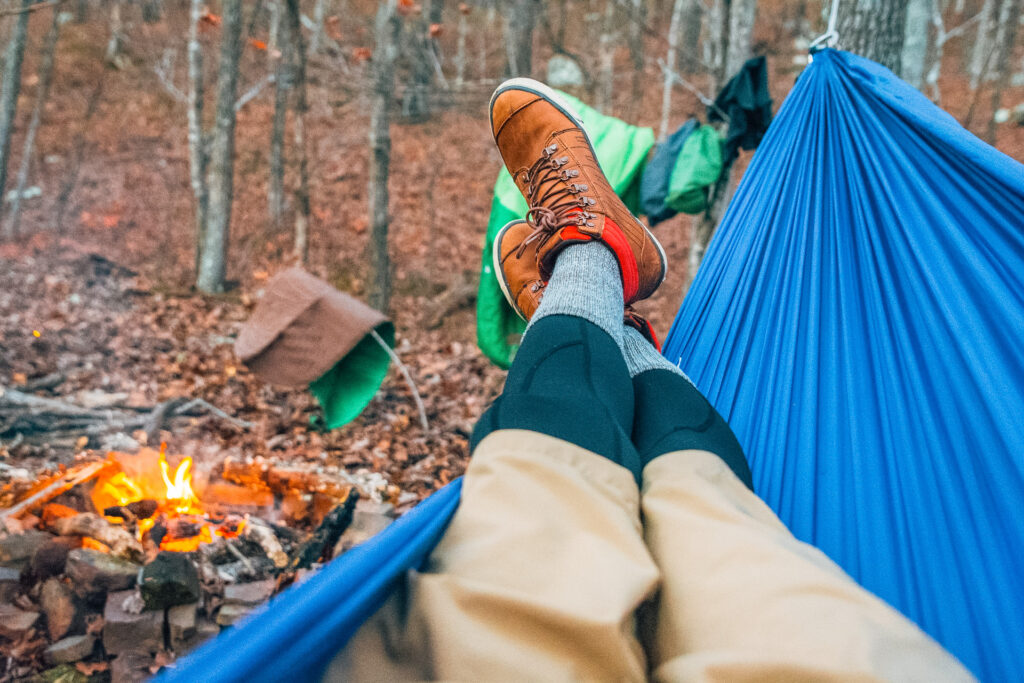 A person relaxing in a camping hammock by a fire in the wilderness.