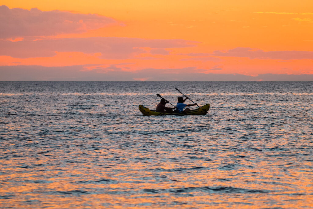 Two people kayaking on the open water as the sun sets and turns the sky orange and pink.