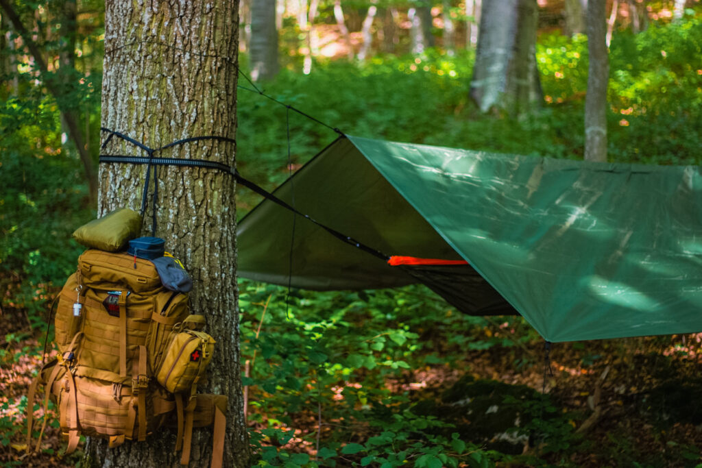 A camping hammock set up under a rainfly in the wilderness surrounded by greenery and trees.