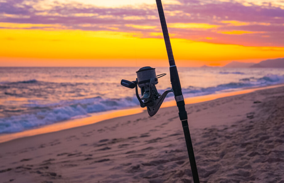 A surf fishing rod set up on the beach as the sun sets in the distance and makes the sky turn yellow, orange, and pink. 