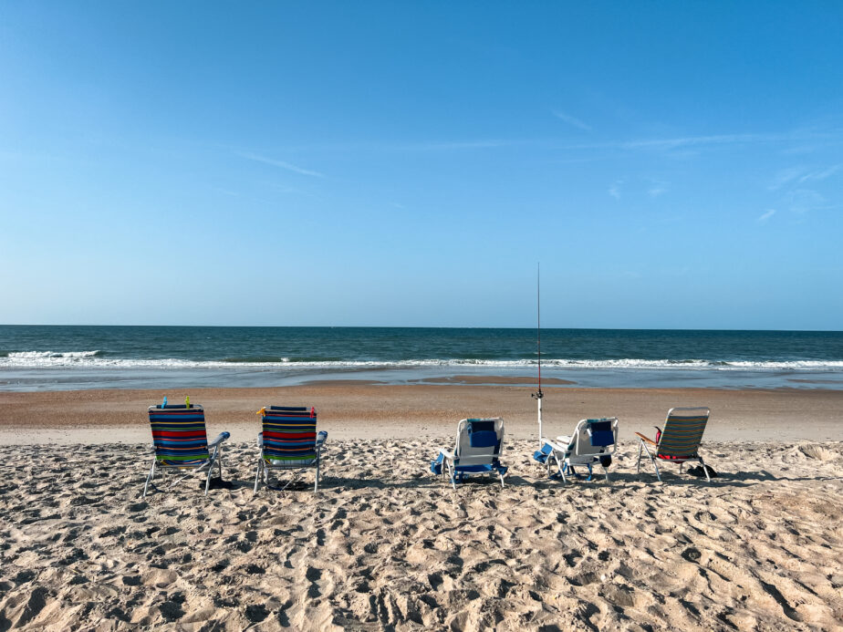 Five chairs set out on the sand with a surf fishing rod and the ocean in the distance.