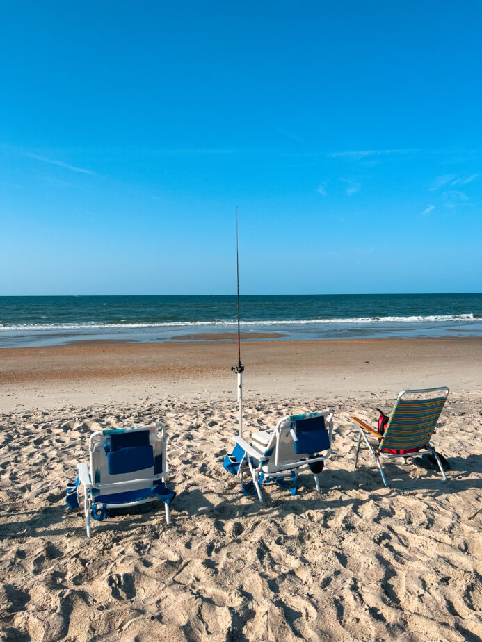 Three chairs set up on the beach with a surf fishing rod in its holder.