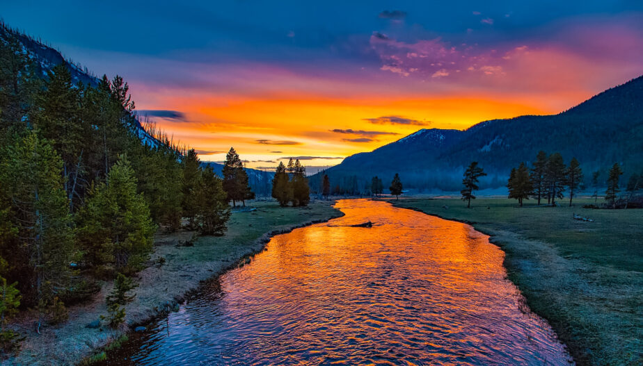 A river running through Montana with mountains in the distance and the sun setting in the background.