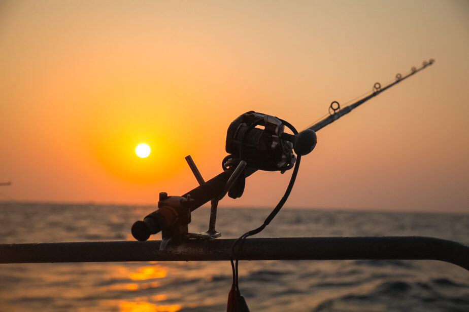 A surf fishing rod and reel set up on a boat overlooking the ocean as the sun begins to set in the distance.