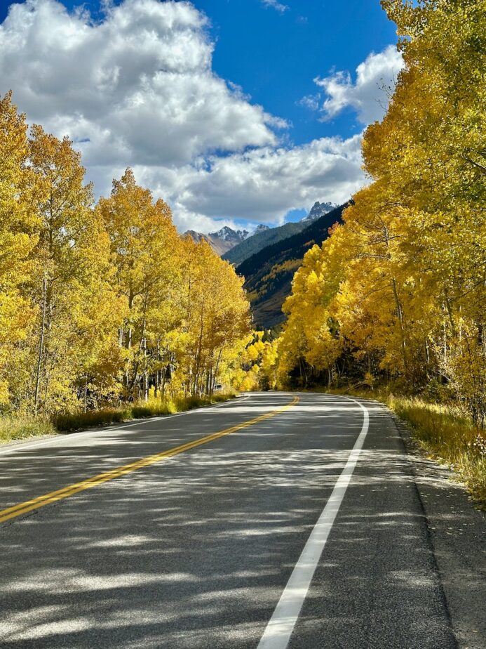 A road in Colorado surrounded by fall foliage with mountains in the distance.