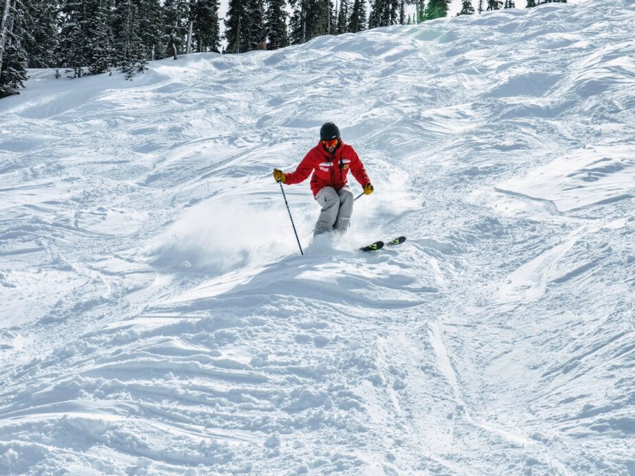 A skier in a red jacket going down moguls at a ski resort.
