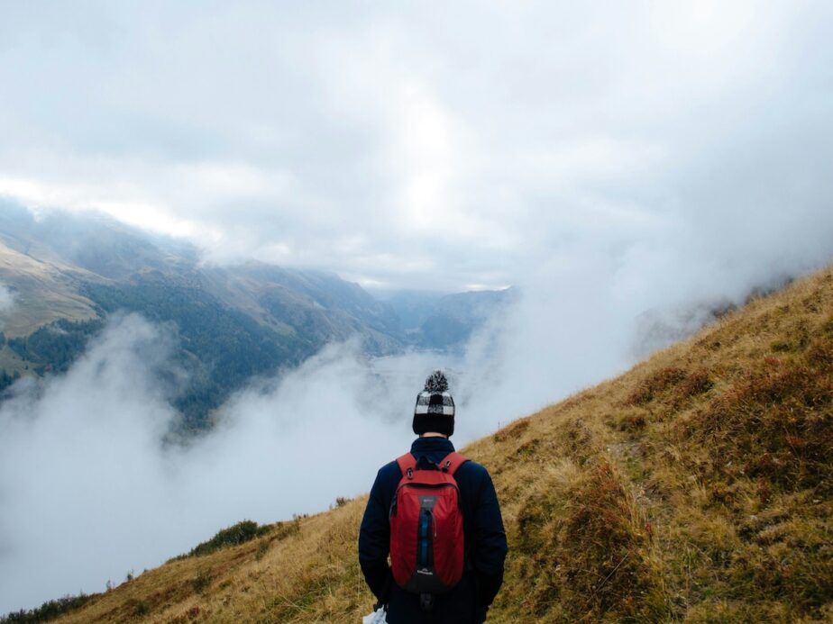 A hiker trekking along grass with clouds and mountains in the distance.